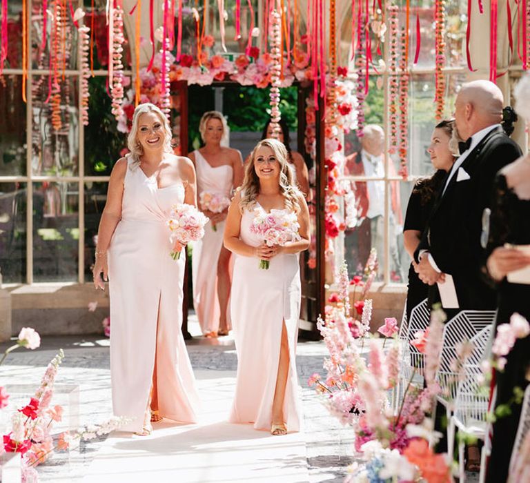 Bridesmaids walk down the aisle in pairs in one shoulder pink bridesmaid dresses 