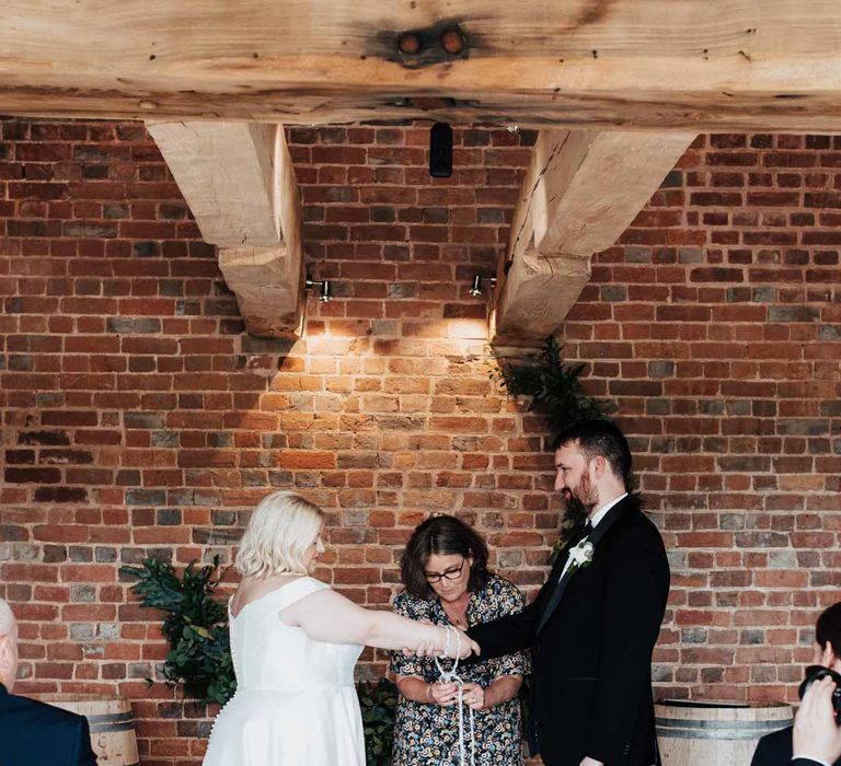 Bride in satin off the shoulder wedding dress partaking in wedding handfasting ceremony at Brickhouse Vineyard Devon wedding with groom in classic black grooms three piece tuxedo with white rose boutonniere and black bowtie 