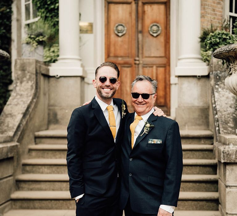 Groom and father of the groom in matching navy suits with yellow ties wearing sunglasses 