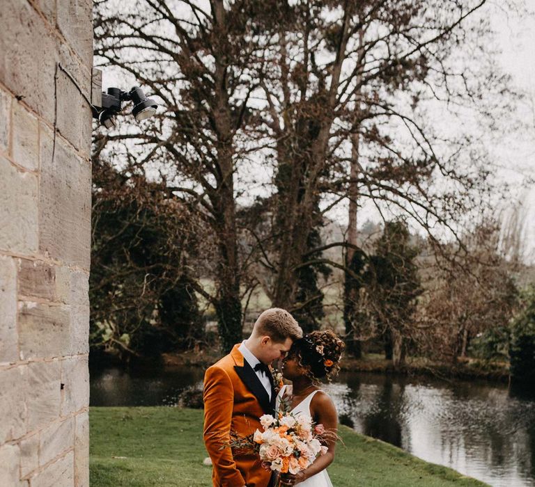 Groom in crushed burnt orange velvet grooms blazer, black bowtie and black suit trousers embracing bride in v-neck sleeveless wedding dress with 3D floral applique train holding blush and orange garden roses, poppies and carnations, dried flowers and eucalyptus wedding bouquet at Brinsop Court wedding venue