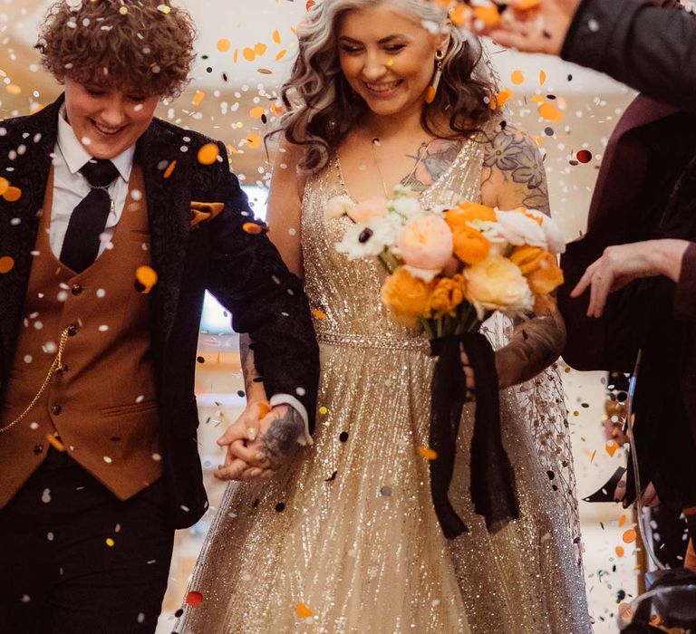 Bride in sleeveless deep v-neck sparkly gold wedding dress with sheer and gold polka dot wedding veil holding orange garden rose, white poppy, light pink carnation and foliage bridal bouquet wrapped in sparkly gold ribbon doing colourful wedding confetti exit with bride in dark velvet bridal jacket with rust waistcoat and pocket square, black suit trousers and gold pocket watch 