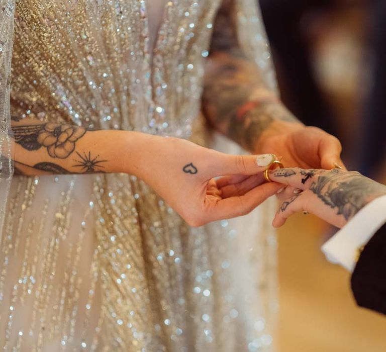 Bride in sparkly gold wedding dress giving bride golden wedding ring at Kingscote Barn Tetbury 