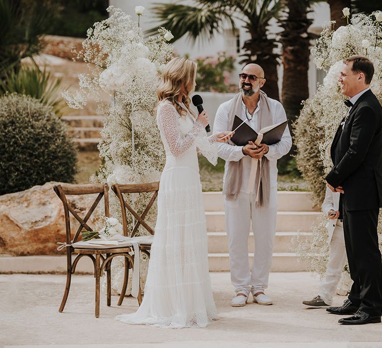 bride in a long sleeve lace wedding dress reading her wedding vows to her groom in a tuxedo at outdoor pure house ibiza wedding ceremony