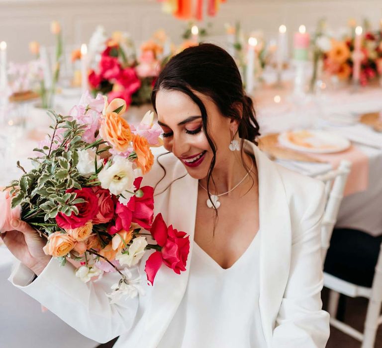 Bride in white bridal suit with silver bridal jewellery holding mixed seasonal bridal bouquet sitting in by wedding tablescape with white wedding tablecloth, burlap place mats, tapered candles, pink and orange wedding stationery, and floral wedding table centrepieces with pink and orange garden roses, orange floribunda, yellow ranunculus flowers, foliage and Iceland poppies with red, pink orange and yellow balloons and wedding streamers at Merriscourt wedding venue