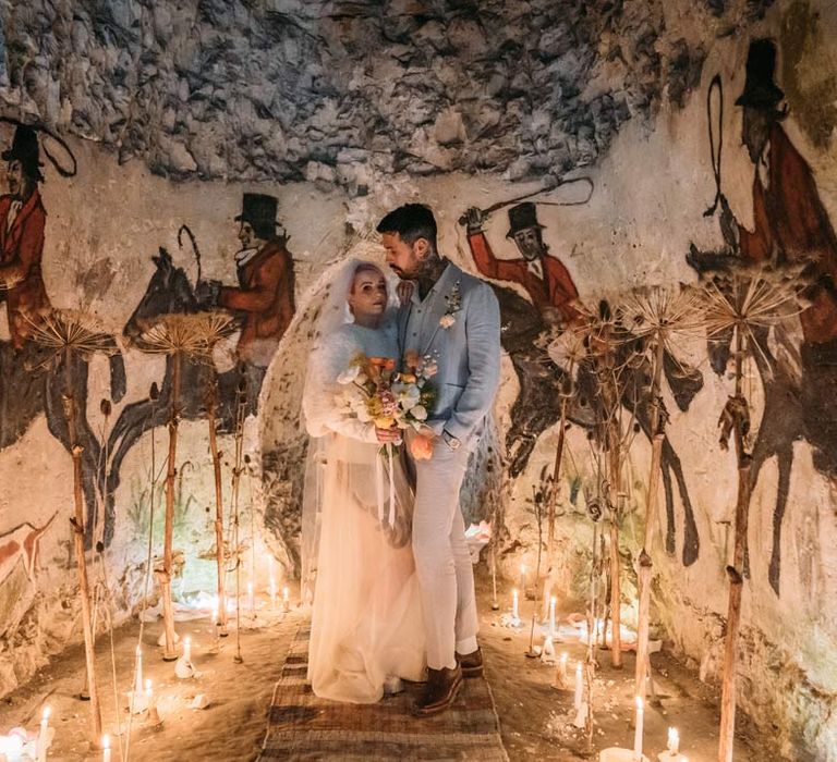 Bride in sheer overlay wedding dress and fluffy bridal coverup standing with groom in baby blue linen suit, personalised embroidered linen shirt and garden rose and pampas grass boutonniere standing around the drawings of Margate Caves