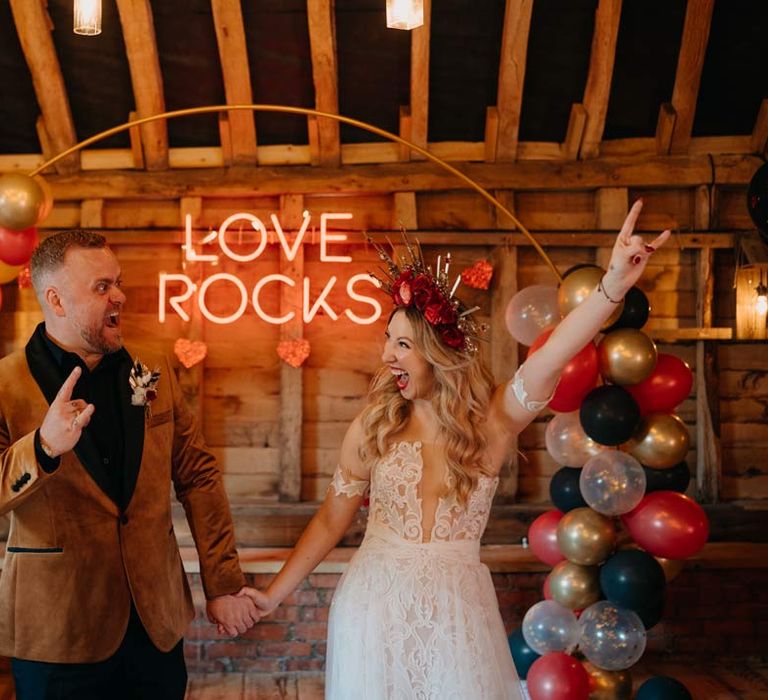 Bride in off shoulder lace wedding dress with corset style top and red rose and gold halo bridal crown holding hands with groom in crushed velvet burnt orange grooms blazer and dried flower boutonniere laughing and posing standing in Southlands Barn wedding venue reception room with a balloon wedding arch and a large pink wedding neon light