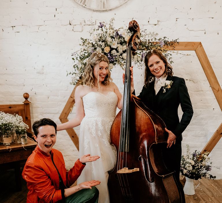 two brides in a navy suit and strapless lace wedding dress posing with their cellist wedding musician 