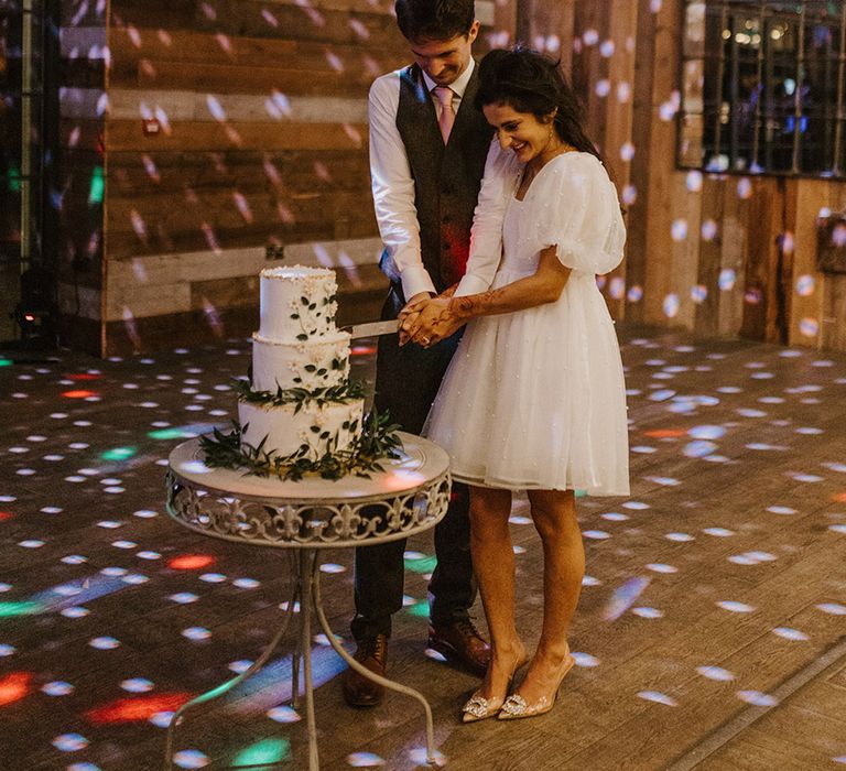 Bride in short puff sleeve reception dress cuts the wedding cake with the groom 