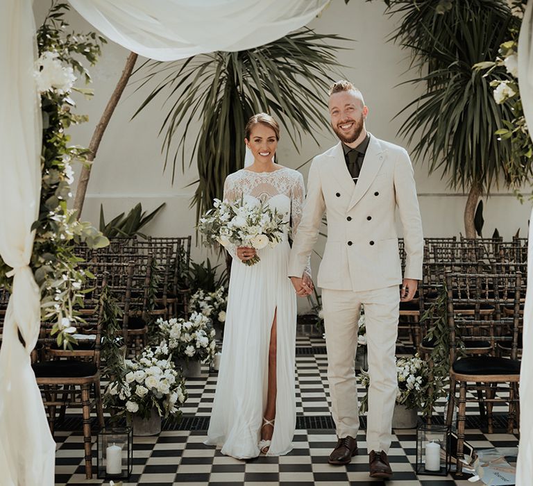 groom in a double breasted beige wedding suit holding hands with his bride in a. Catherine Deane wedding dress under a floral drape altar at Gunnersbury Park 