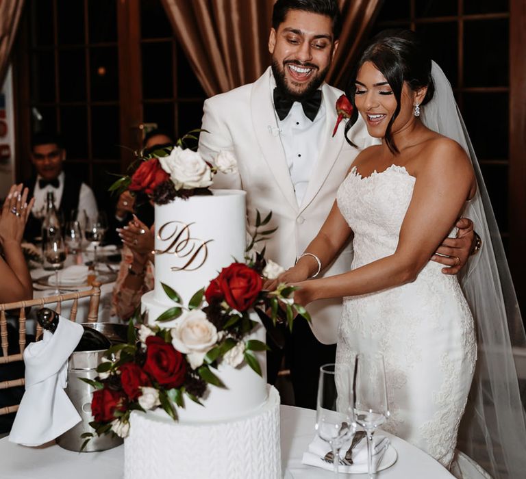 The bride and groom cut their three tier white iced wedding cake with red rose decorations 