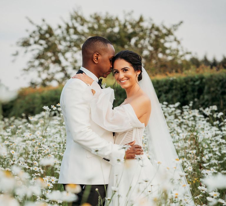 bride in white off-the-shoulder tulle wedding dress with groom in black-tie tuxedo pose for a couple portrait in a daisy garden