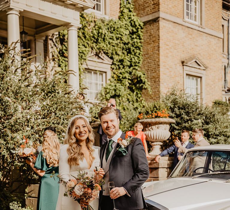 Bride wearing a long sleeve wedding dress with plunging neckline smiling with the groom next to their light blue wedding car 