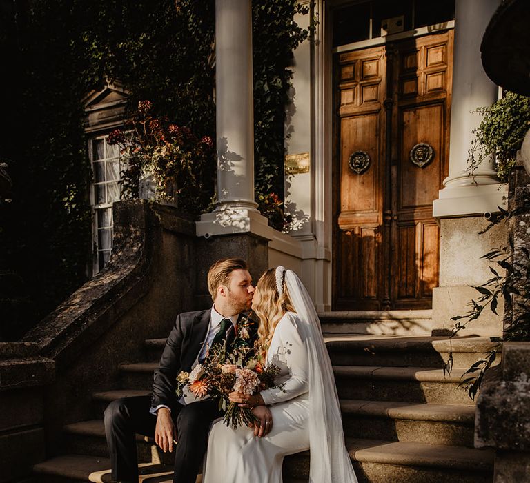 Country house wedding with bride and groom kissing as they sit on the steed of the wedding venue