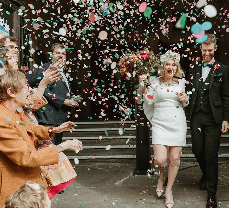 Bride in short square neck wedding dress with groom in black suit and green tie as they walk out of their ceremony to colourful confetti 