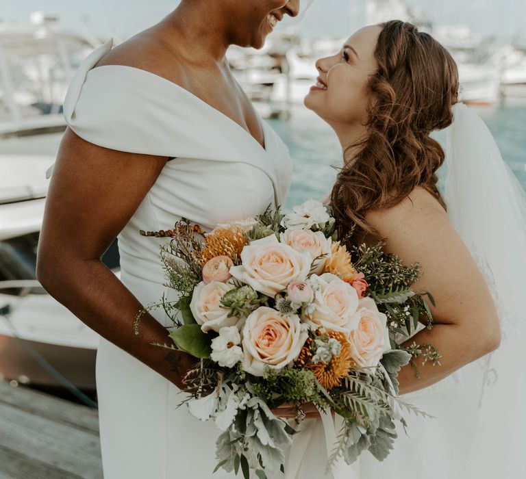 Black bride in a jumpsuit holding the wedding coral, pink, orange and peach bouquet with her bride in a strapless wedding dress 