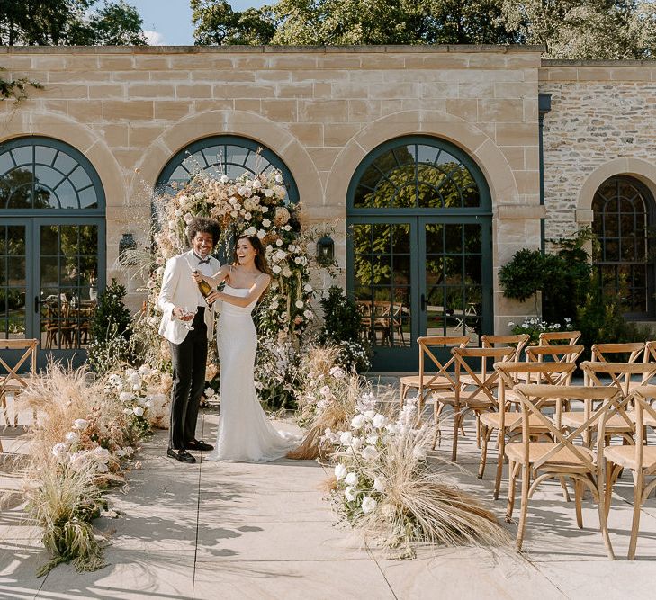 Bride in a sequin wedding dress popping champagne in front of a flower arch at Middleton Lodge 