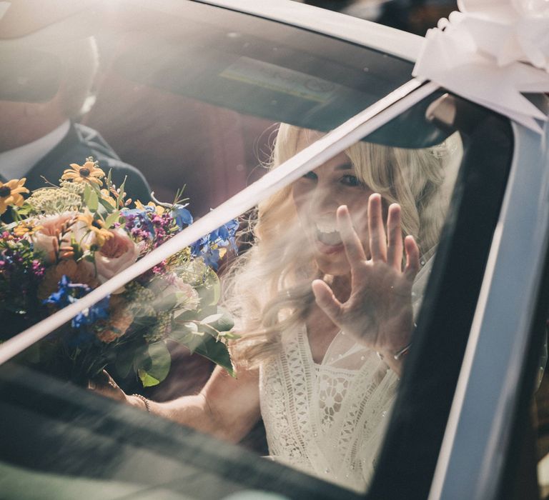 Smiling bride holding colourful bouquet waves from open top car 