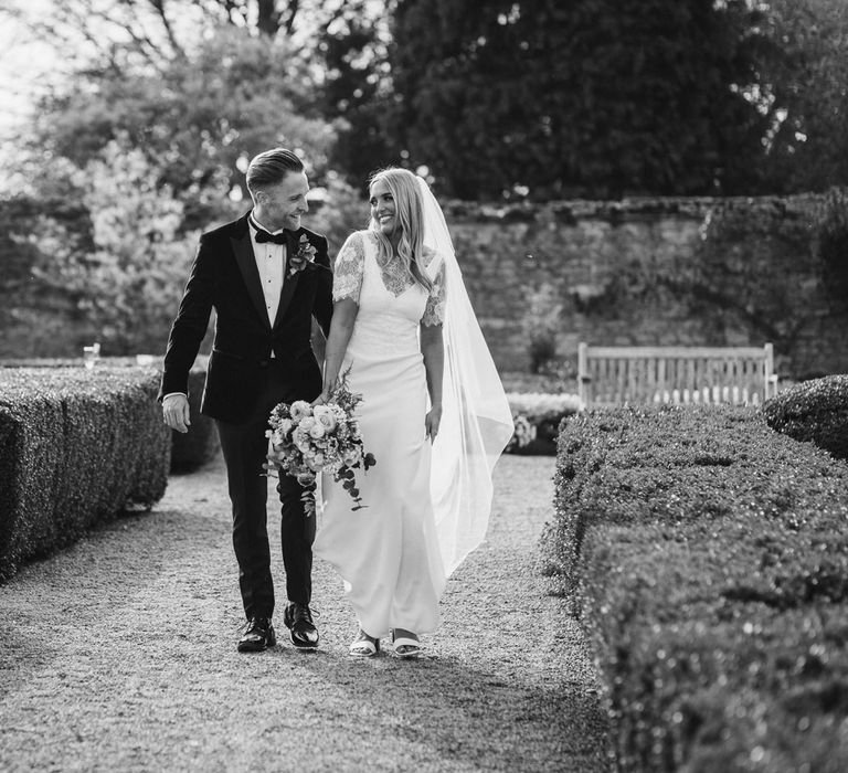 Bride in white Charlie Brear dress, white lace Augusta Jones top and full length veil walking with groom in black velvet Hugo Boss suit in the grounds at Caswell House wedding