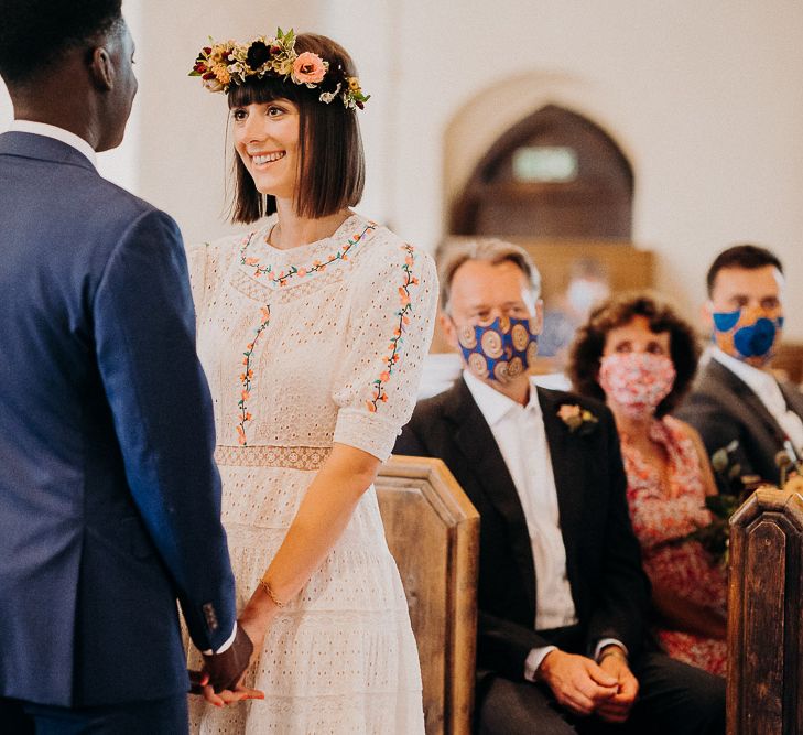 Bride in a Broderie Anglaise wedding dress with colourful embroidery detail holding hands with her groom at the altar 