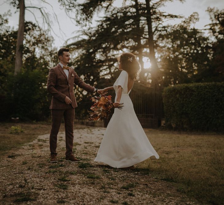 Groom in a brown wedding suit dancing at dusk with his bride in an a-line wedding dress with cap sleeves and button detail