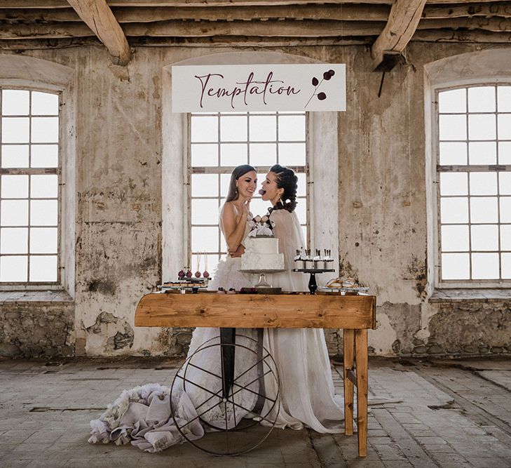 Two brides standing at the dessert cart with individual treats and a two tier wedding cake 