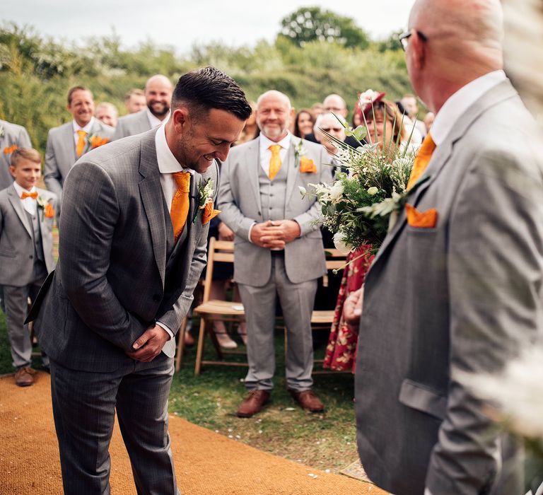 Groom stands laughing at the end of the aisle with father-of the bride and groomsmen all in matching grey Hugo Boss suits, waistcoats and yellow ties