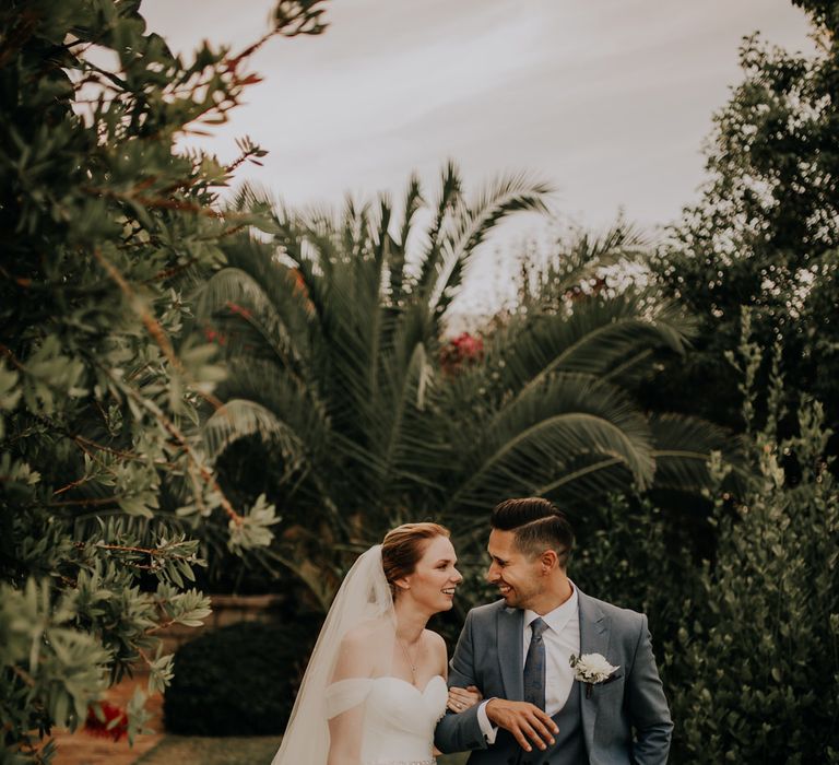 Bride and groom walking arm in arm through lush garden greenery with palm tree in the background