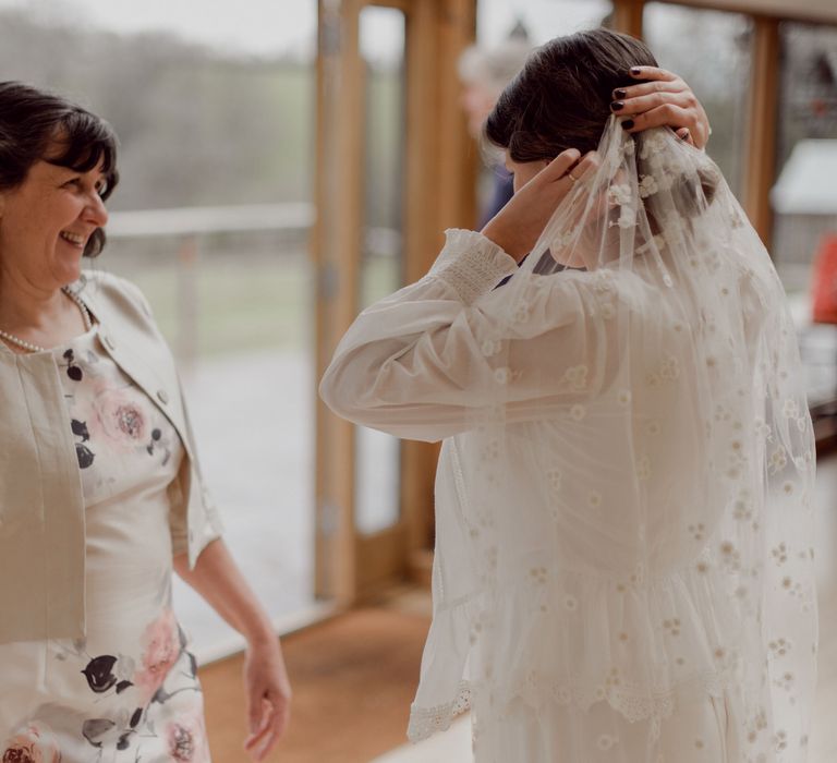 Bride in Charlie Brear wedding dress and white blouse adjusts daisy applique veil opposite smiling wedding guest in white floral dress