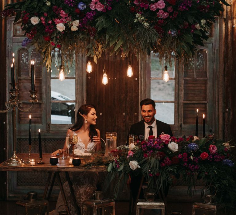 Bride and groom sitting underneath moody romantic floral installation with Edison bulbs and pillar candles