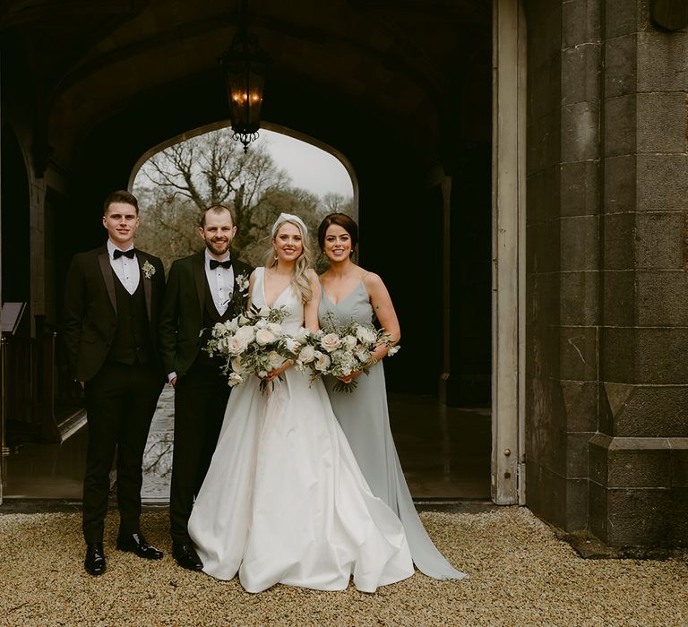 Bride & groom stand beneath arch outdoors whilst stood with wedding party 