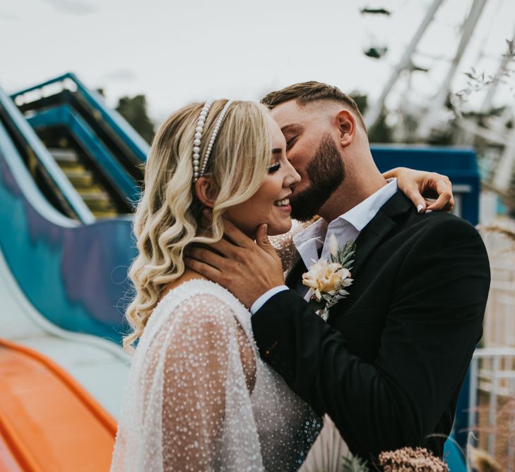 Groom in a open collar white shirt and black suit kissing his bride in a sparkly wedding dress with pearl headband at Dreamland Margate