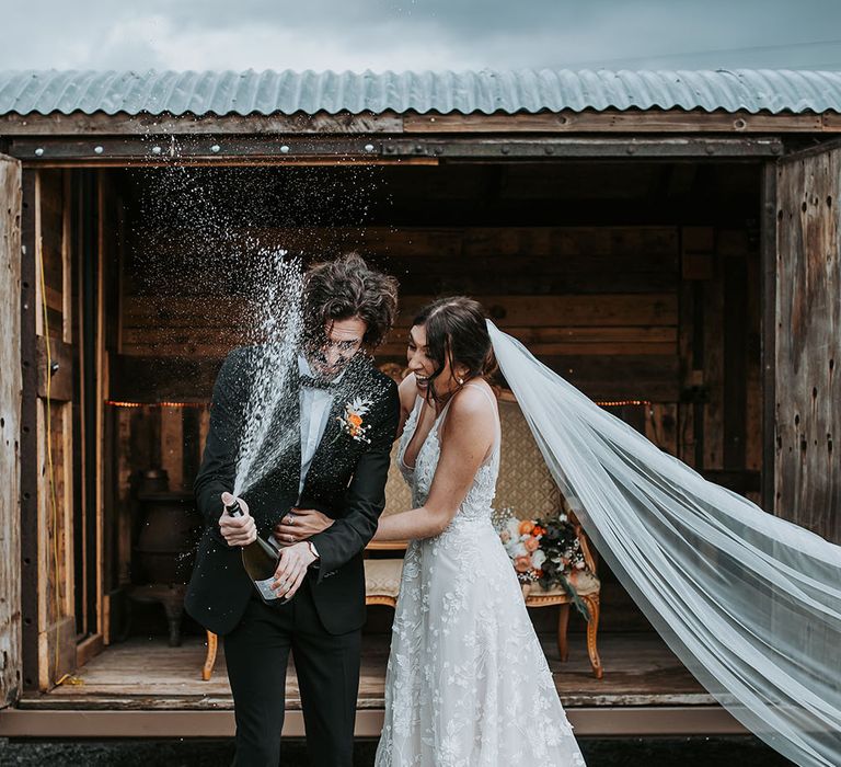 Groom in a black tuxedo popping champagne with his bride in an appliqué wedding dress at new Lake District wedding venue, Ghyll Barn 