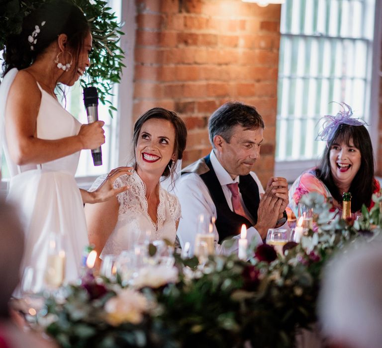 Bride in white cami wedding dress holding microphone rests hand on shoulder of bride in white lace top capped sleeve wedding dress as they smile at each other during wedding speech at The West Mill Derby