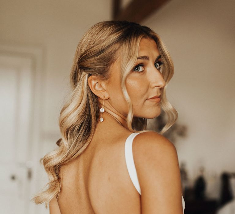 Bride with curled blond hair wearing open back Made With Love wedding dress and gold drop earrings looks over her shoulder before wedding at Anran Devon