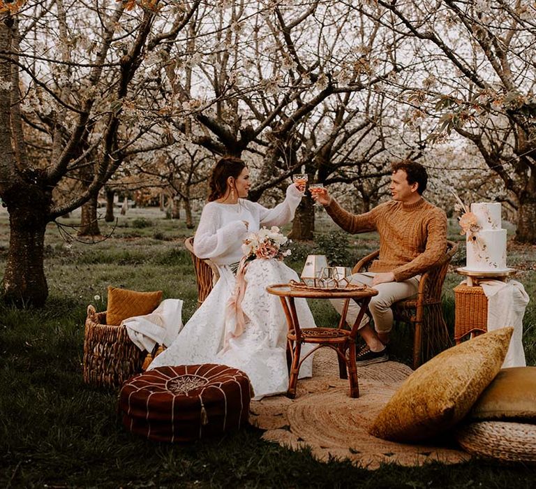 Bride and groom in jumpers toasting in the forest sitting on wicker chairs 