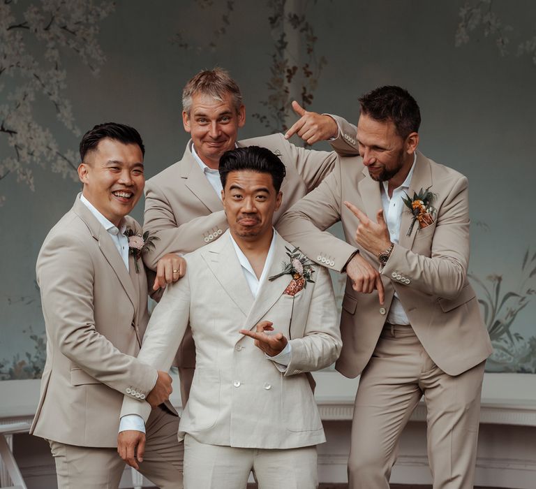 Groom in white shawl collared shirt and double breasted linen suit stands outside Victorian summerhouse with groomsmen in white shirts and linen suits all pointing at each other for Wasing Park wedding