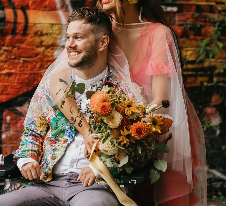 Bride in a coloured wedding dress with cape veil embracing her disabled groom in grey chinos and brightly coloured jacket 