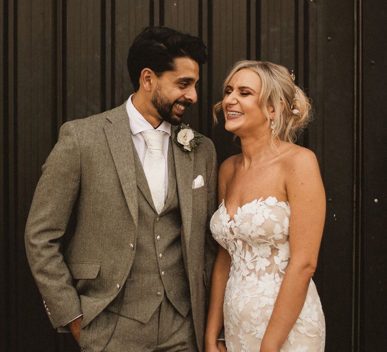 Groom in grey three piece suit with white tie and floral buttonhole smiles at laughing bride in white off the shoulder Enzoani wedding dress at Inkersall Grange Farm wedding