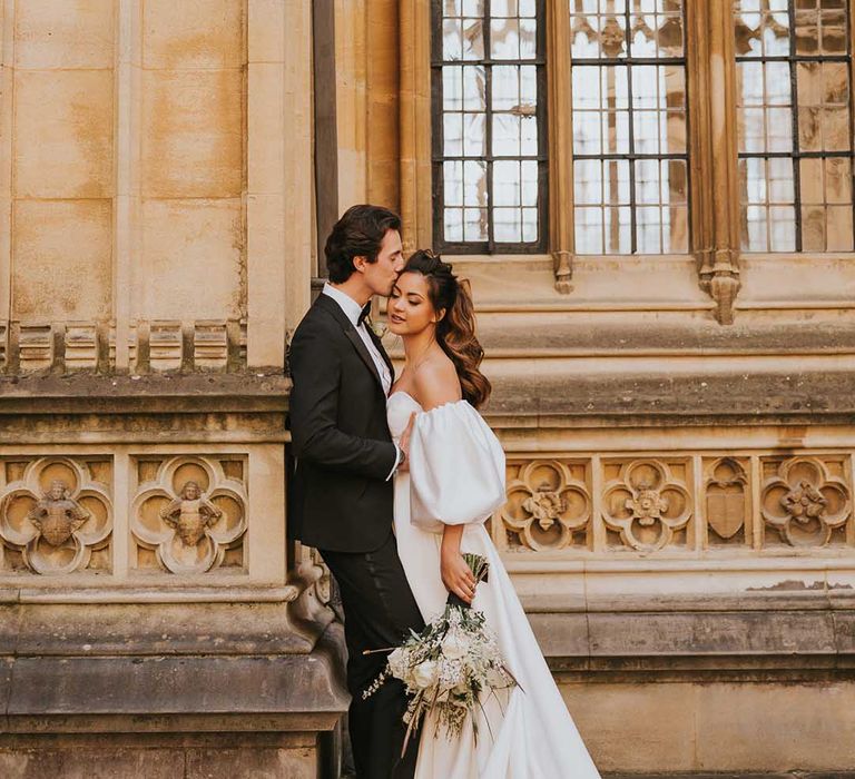 Groom in a tuxedo kissing his bride in a strapless wedding dress with puff sleeves