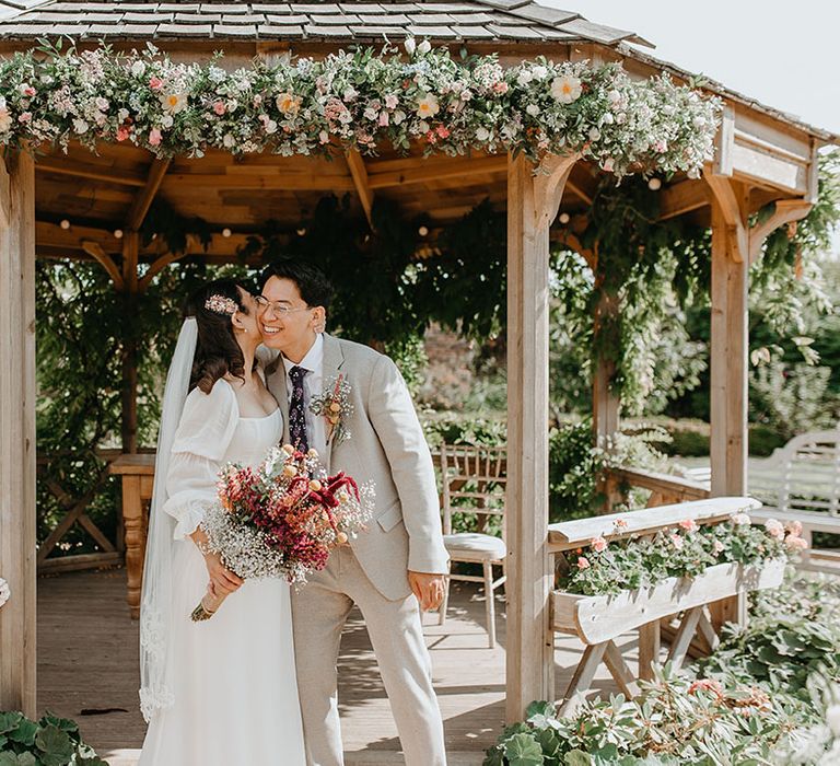 Bride & groom kiss in front of pergola covered in pastel florals