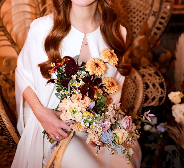 Smiling bride in white cape sits on large rattan chair holding mutilcoloured pastel bridal bouquet 