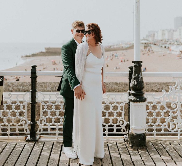 Groom stands behind groom and wraps his arms around bride on their wedding day in front of Brighton beach front