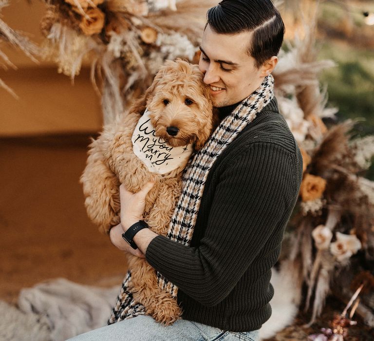 Groom-to-be in a black jumper and check scarf holding his pet cockapoo with a bandanna 