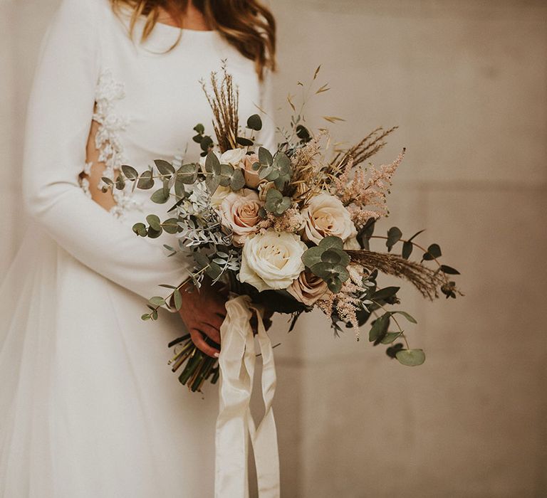 Bride in a long sleeve wedding dress holding a romantic pink rose and green foliage wedding bouquet tied with ribbon