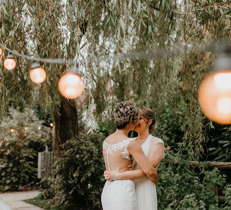 Brides kiss outdoors under festoon lighting next to green foliage