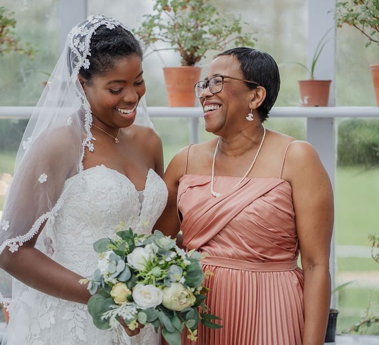 Mother of brides smile in pink toned pleated dress as she cuddles her daughter in a stunning lace veil & simple beautiful make up