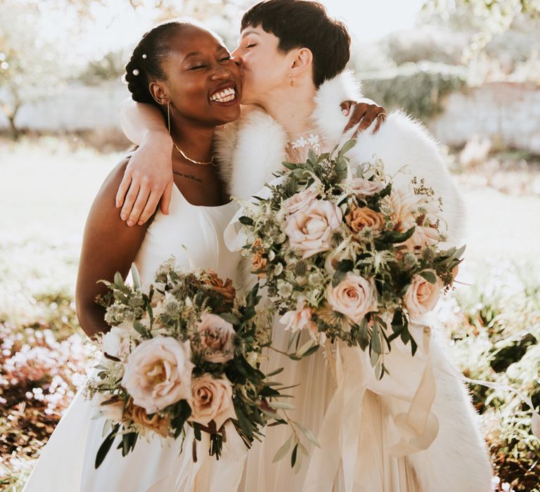 Bride with short brown hair kissing her Black bride with pearl hair accessories in her hair holding matching pink and green wedding bouquets 