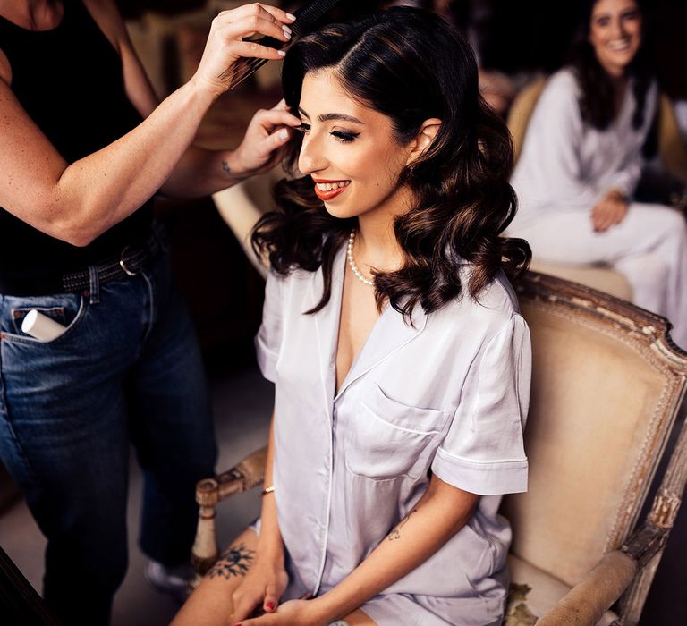 Bride in short sleeve pyjamas wearing pearl necklace has her hair done before wedding