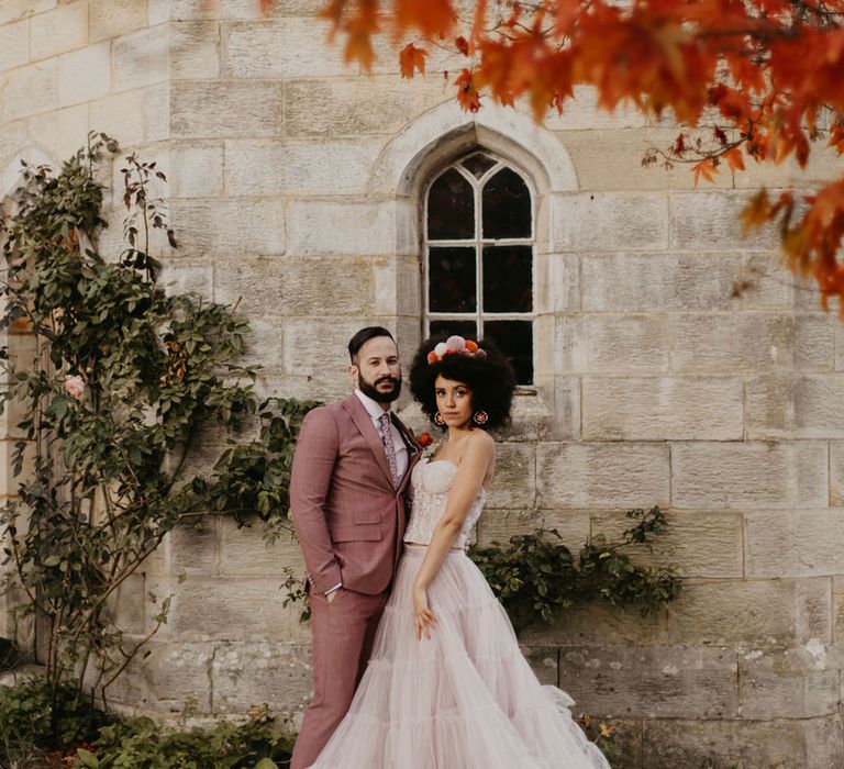 Black bride with afro hair wearing a pompom headband and a pink tulle skirt and strapless corset top with her groom in a pink suit 