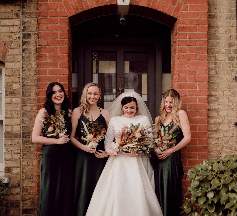 Bride in long sleeve high neck pleated wedding dress and pearl headband with bridesmaids in forest green satin dresses in front of house